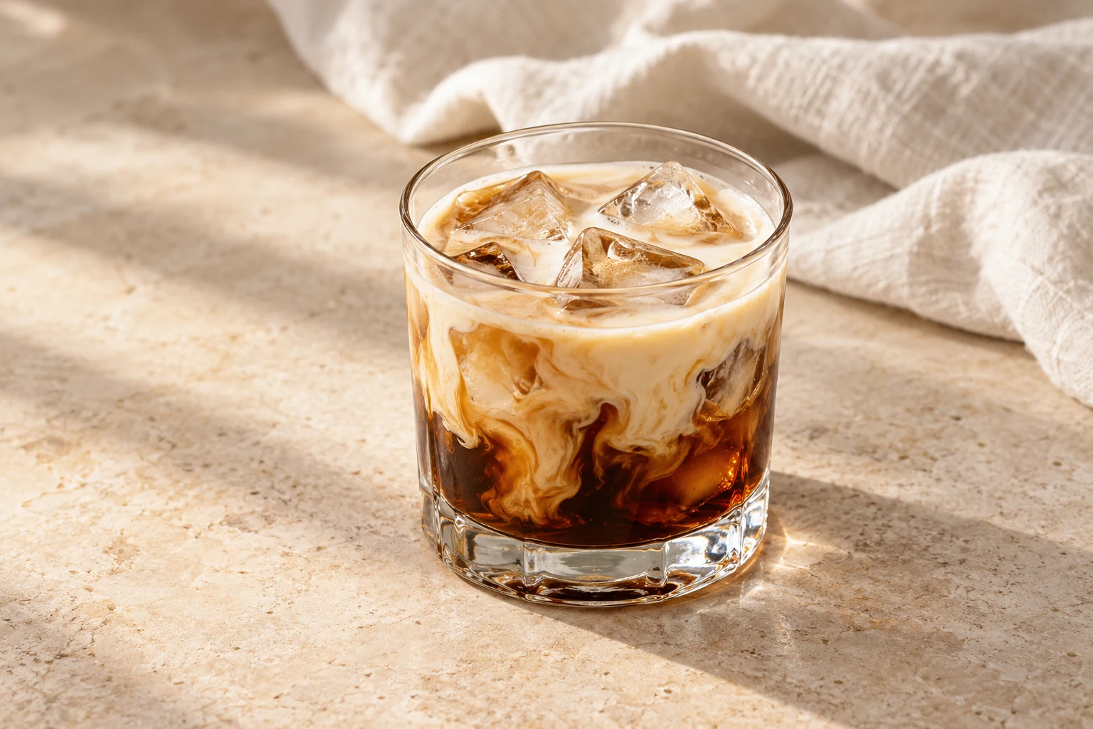 Coffee-brown White Russian over irregular ice in a short rocks glass, with pale cream drifting through the drink and no garnish